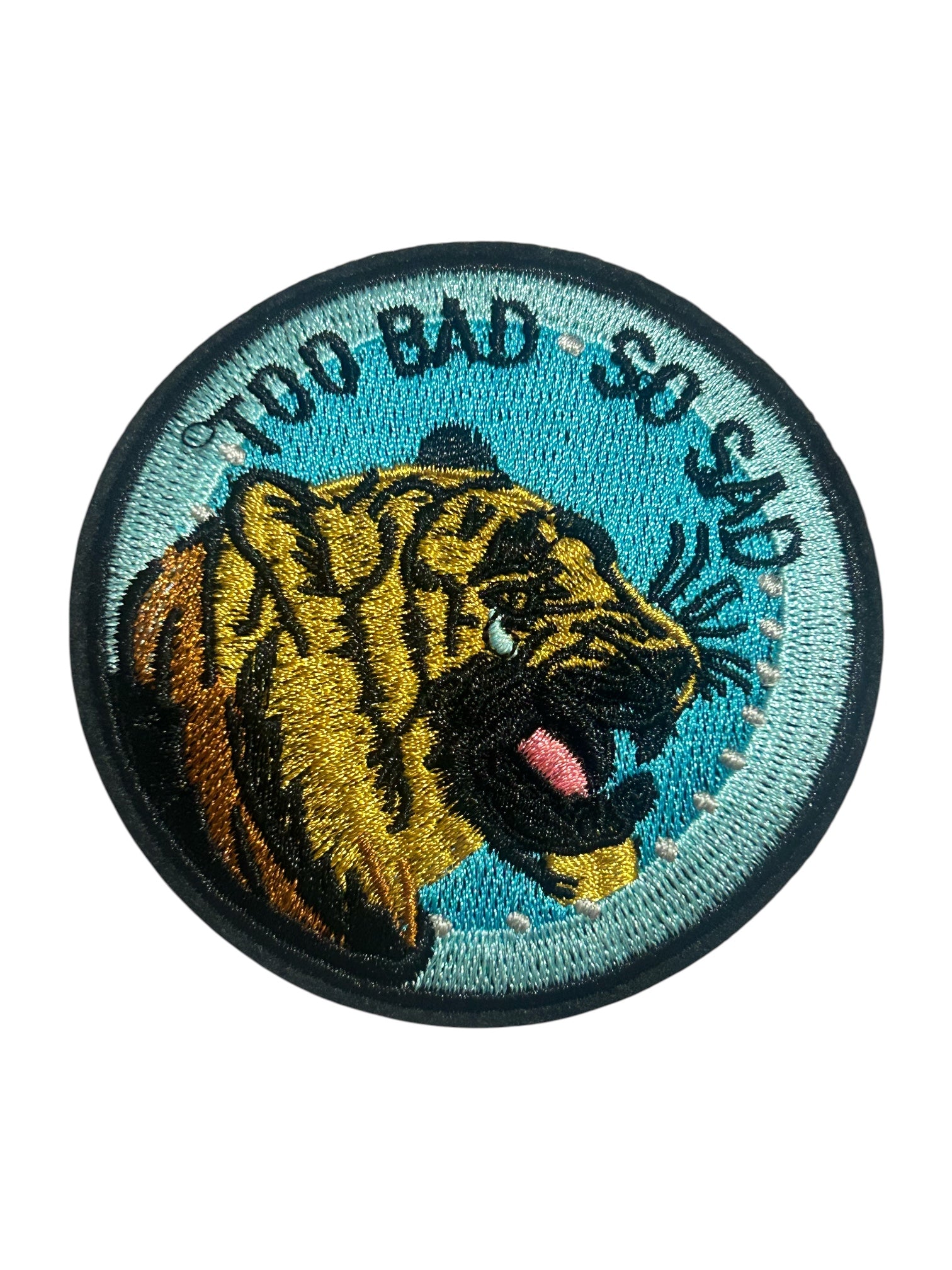 A circular embroidered patch featuring a roaring tiger head on a light blue background with the text 'TOO BAD SO SAD' in black around the top arc.