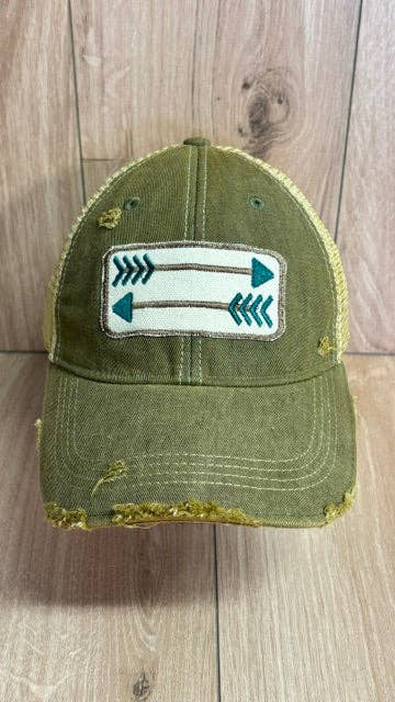 An olive green distressed trucker cap with a white rectangular patch on the front featuring teal arrows, set against a light wood plank background.