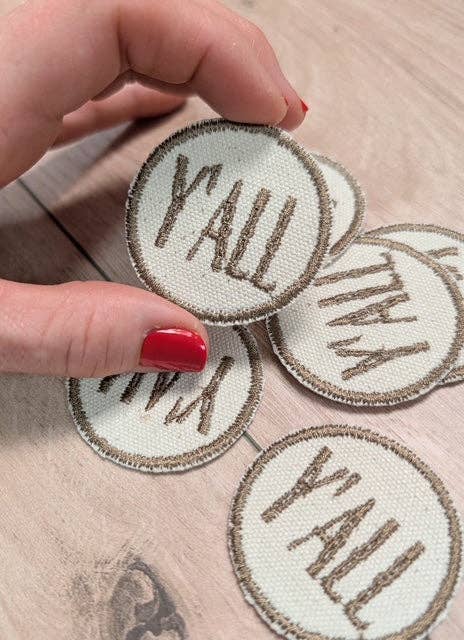 Circular patches with 'Y'ALL' text held by a hand on a wooden surface