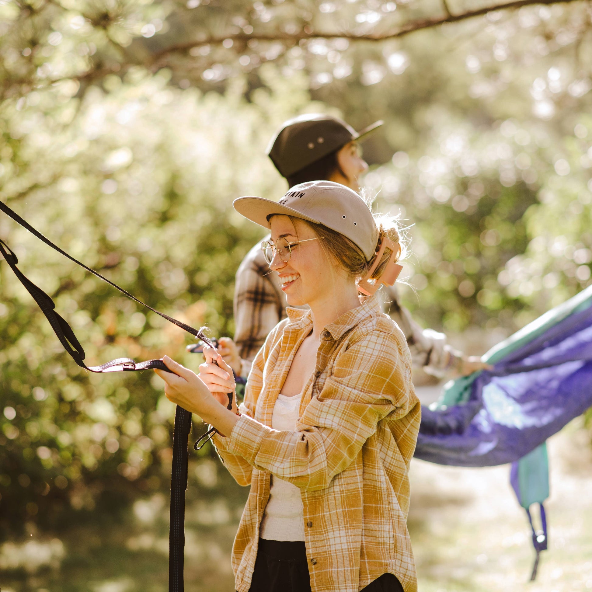 A woman in a yellow plaid flannel shirt and beige cap smiles while holding camping gear outdoors, with another person and a blue hammock visible in a sunlit wooded setting.