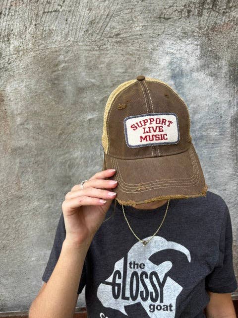 A person wearing a brown distressed trucker hat with a white patch that reads 'SUPPORT LIVE MUSIC' in red, holding the brim in front of their face. They are wearing a dark gray t-shirt with a white graphic and standing against a textured gray concrete wall.