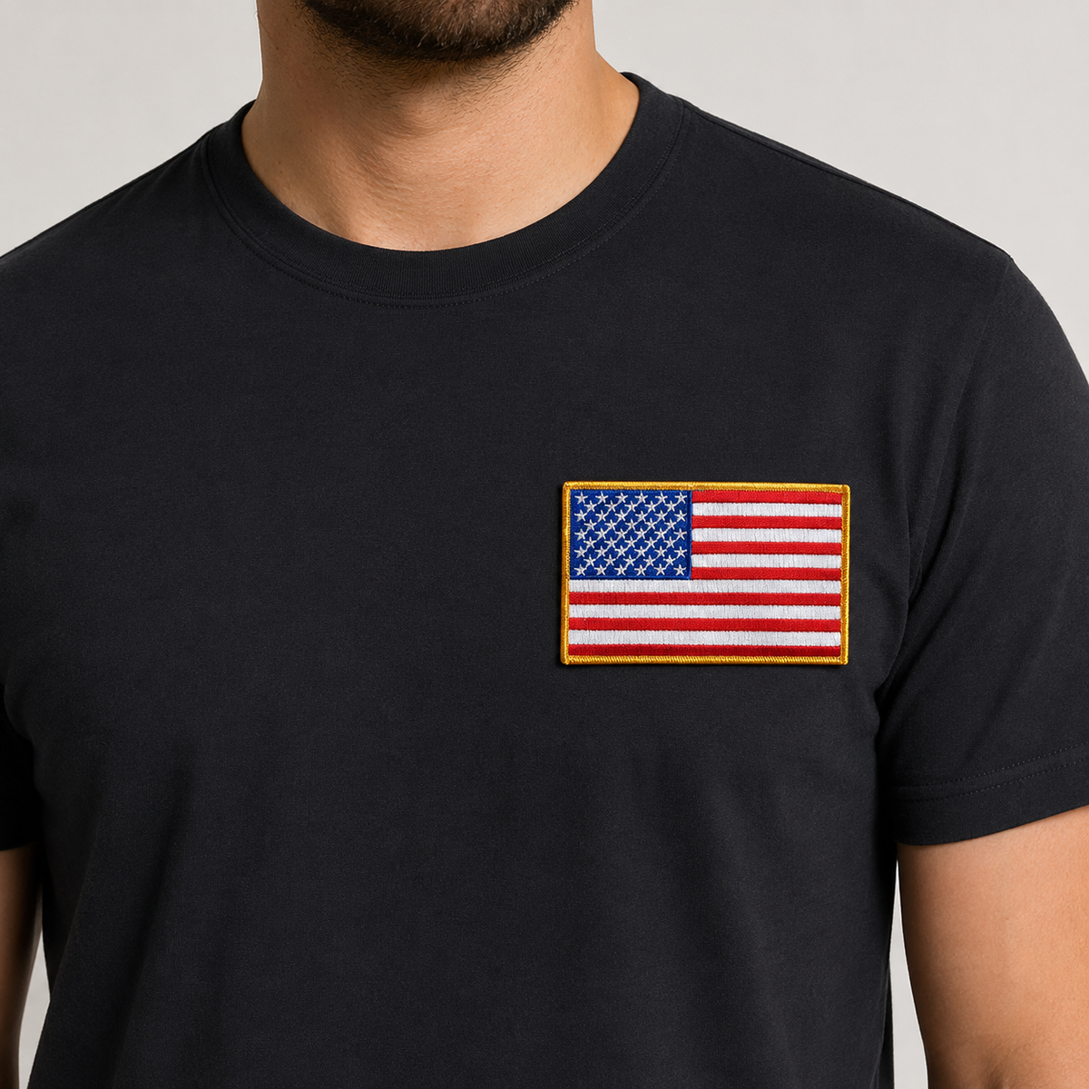 A close-up of a man wearing a black crew-neck t-shirt with a rectangular embroidered American flag patch on the left chest, set against a plain light gray background.
