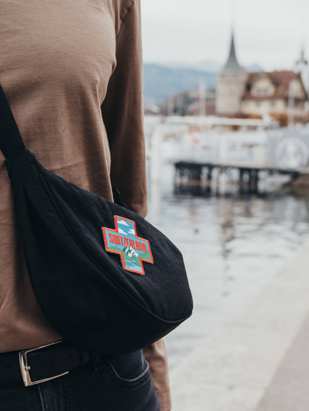 A person wearing a brown shirt and black pants carries a black sling bag with a colorful embroidered 'Swiss Made' patch, set against a blurred waterfront background.
