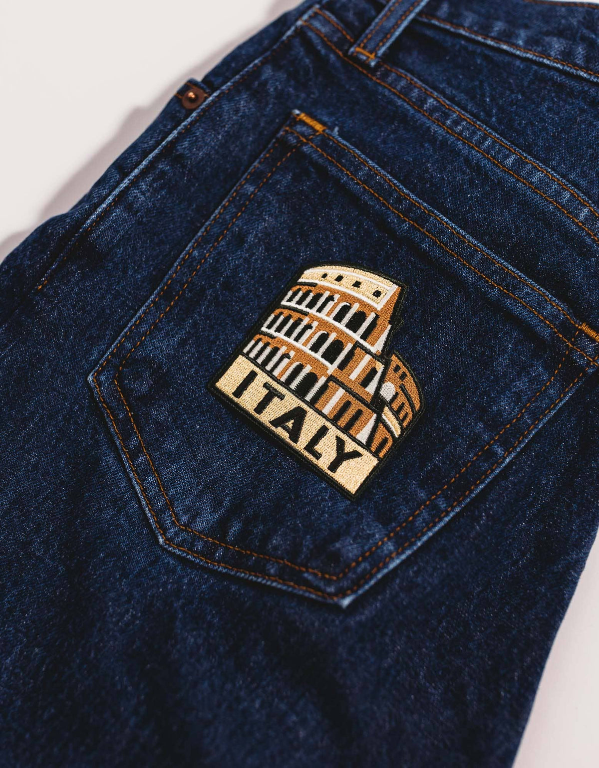 Close-up of a dark blue denim jeans pocket featuring a decorative embroidered patch of the Colosseum with the word 'ITALY' on a light background.