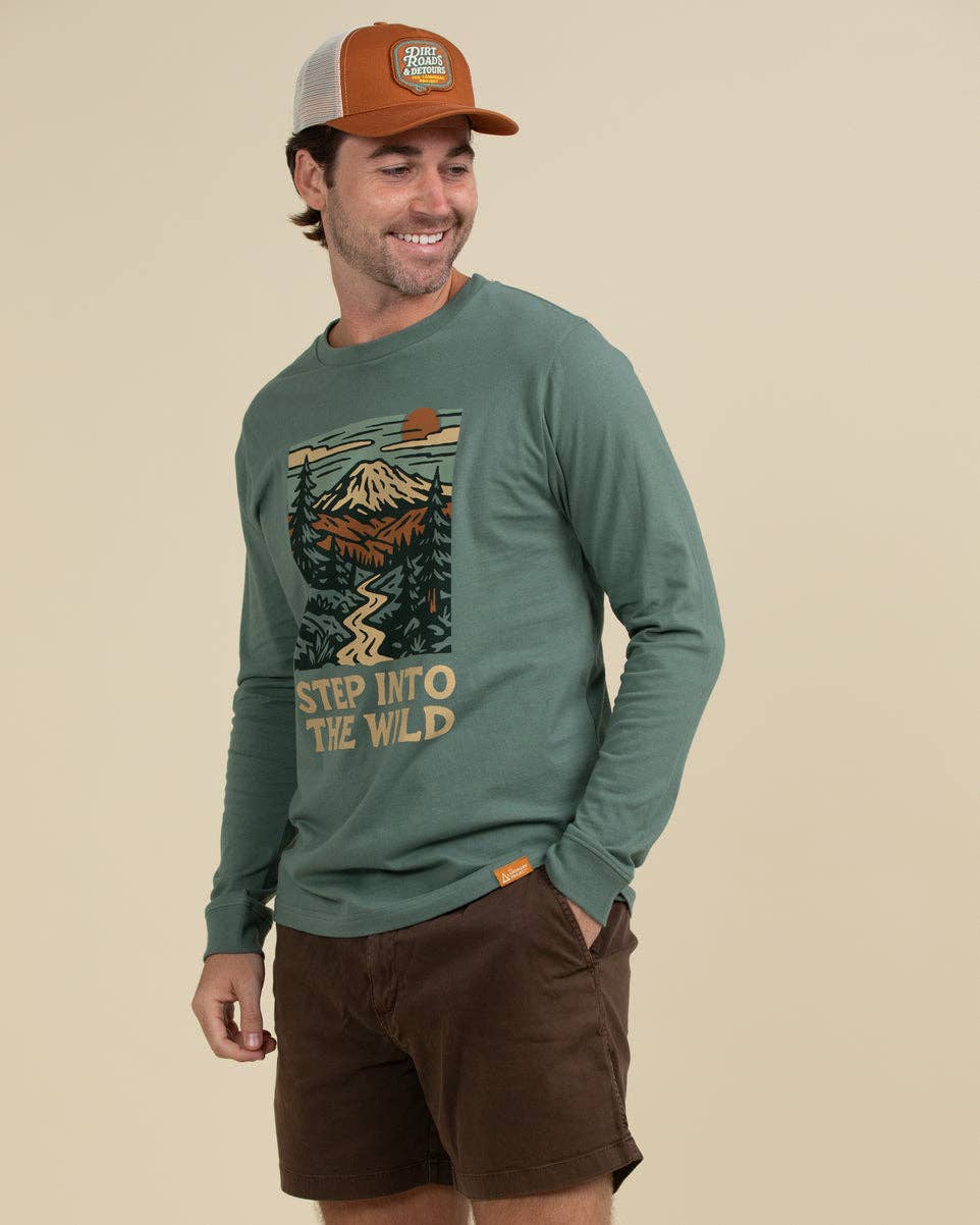 A man wearing a sage green long-sleeve graphic t-shirt with a mountain print and the text 'STEP INTO THE WILD', paired with brown shorts and an orange and gray trucker hat, against a solid beige background.