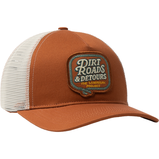 A brown and white trucker hat with a mesh back, featuring a brown embroidered patch on the front that reads 'Dirt Roads & Detours The Landmark Project'.