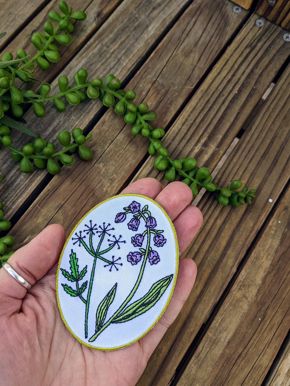 A hand holds an oval embroidered patch featuring purple flowers and green leaves on a white background, with a wooden deck and green trailing plants in the background.