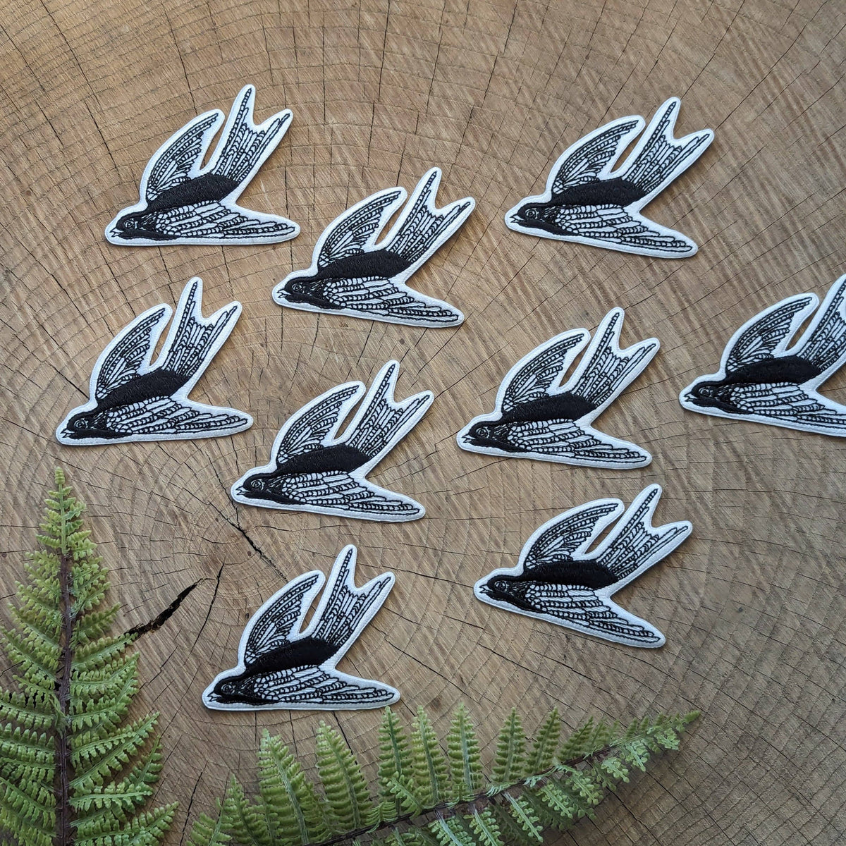 Nine black and white swallow-shaped embroidered patches arranged on a wood slice background with green fern leaves at the bottom.