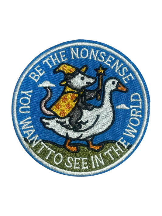 A circular embroidered patch featuring a white goose carrying a gray cat in a yellow robe, with the text 'YOU WANT TO SEE IN THE WORLD BE THE NONSENSE' around the border on a white background.