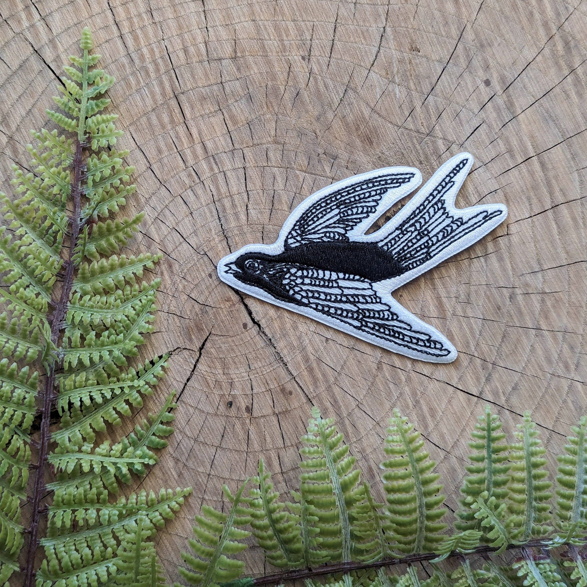 A black and white embroidered patch of a flying swallow bird, styled on a rustic wood slice background with green fern leaves.