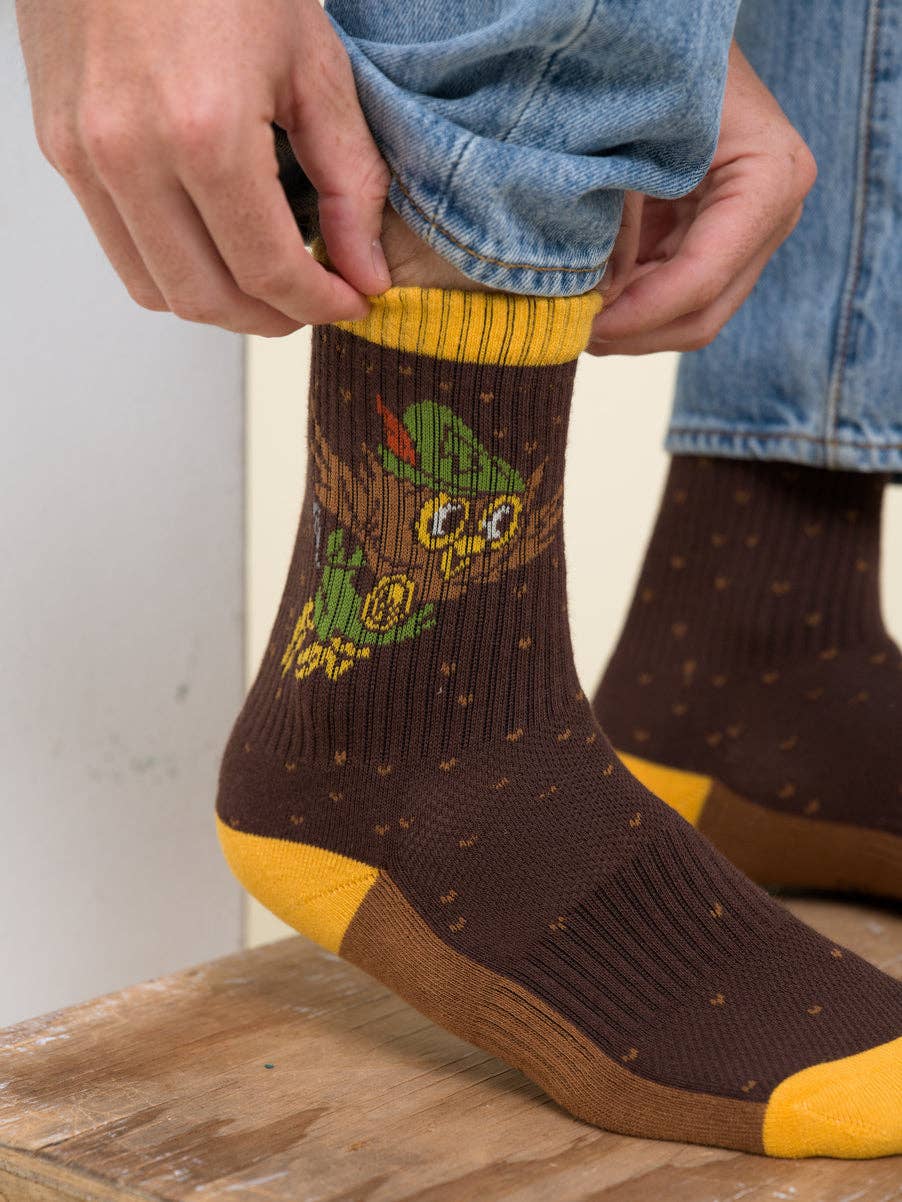 A person wearing brown crew socks with a green Yoda graphic and yellow accents, shown while pulling the top sock up with both hands. Blue jeans and a wooden floor are visible in the background.