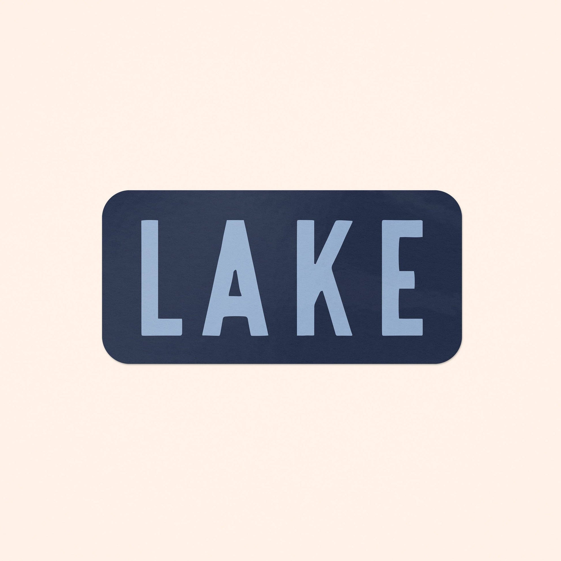 A rectangular sticker with rounded corners featuring the word 'LAKE' in light blue sans-serif letters on a dark navy blue background, centered on a solid pale peach-beige backdrop.