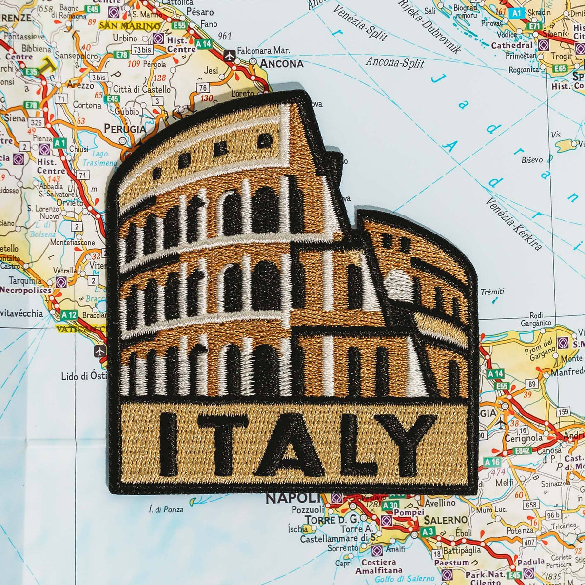 An embroidered patch of the Colosseum in Rome, Italy, featuring a tan and brown architectural design with white accents and the word 'ITALY' at the bottom, displayed on a colorful map background.