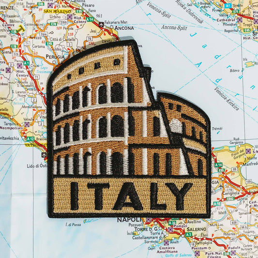 An embroidered patch of the Colosseum in Rome, Italy, featuring a tan and brown architectural design with white accents and the word 'ITALY' at the bottom, displayed on a colorful map background.