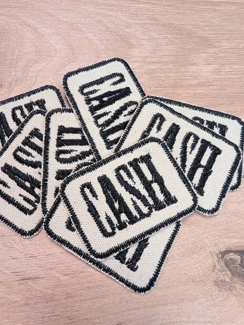A pile of rectangular embroidered patches with rounded corners, featuring the word 'CASH' in bold black lettering on an off-white background with black stitched borders, arranged on a light wood surface.