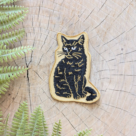 Embroidered patch of a cat on a wooden surface with ferns around