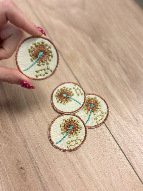 Three embroidered dandelion patches on a wooden surface with a hand holding one patch.