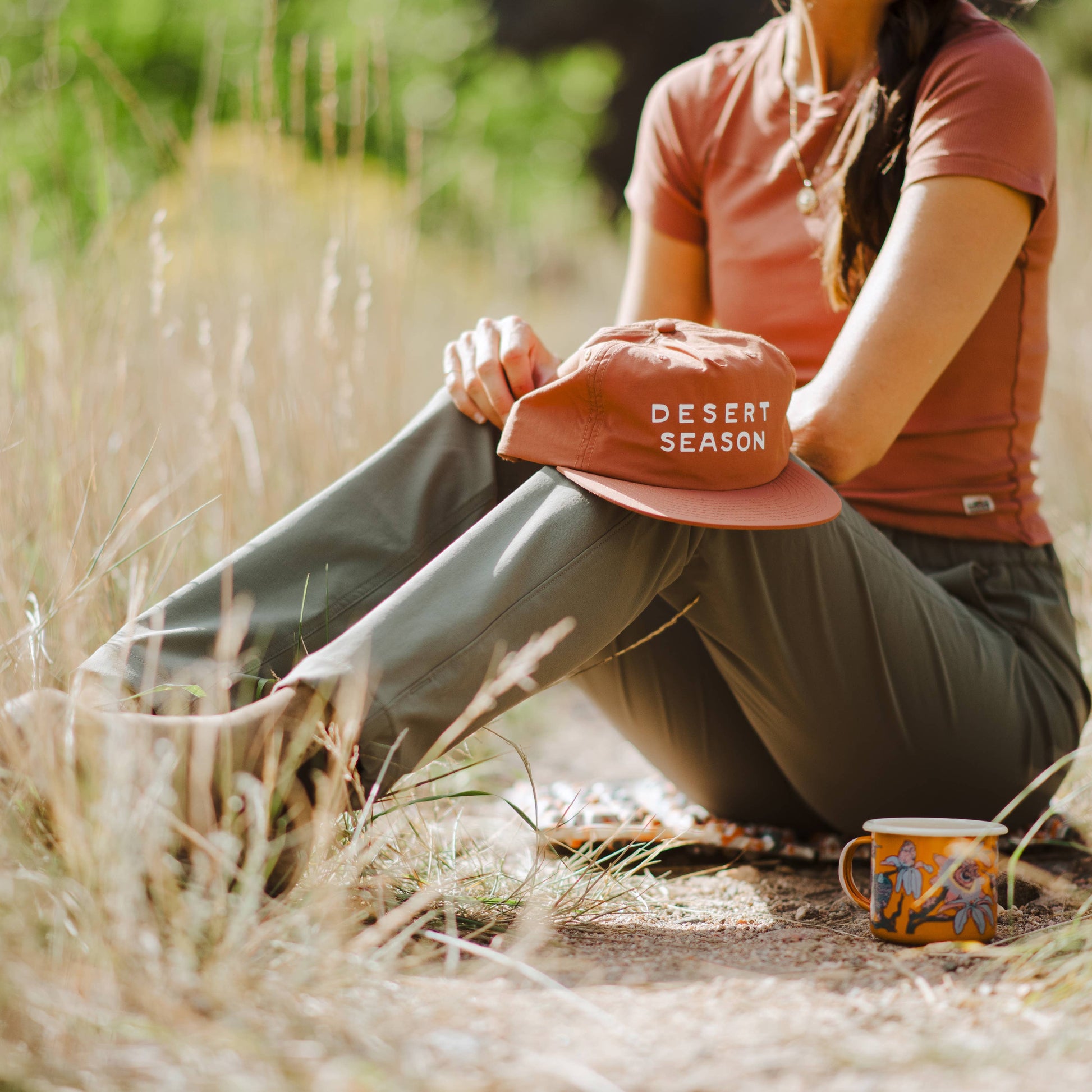 A woman sitting outdoors in dry grass wearing a rust-colored t-shirt, olive green pants, and holding a rust-colored baseball cap that says 'DESERT SEASON' in white text. A patterned mug sits nearby on the ground.