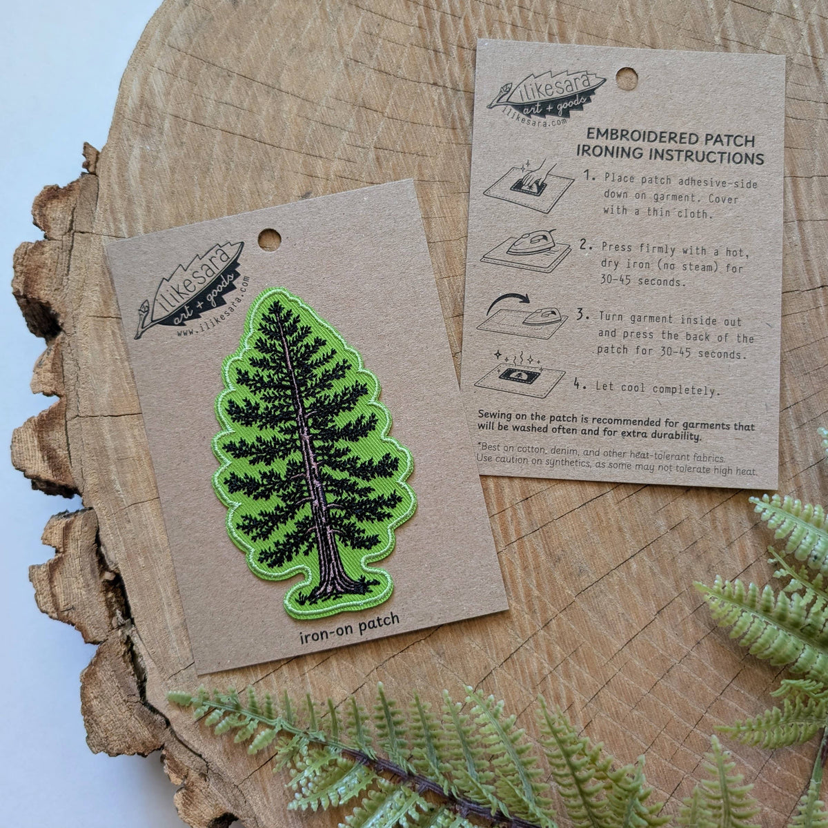 A green embroidered pine tree iron-on patch displayed on a kraft paper backing card, with an instruction sheet and a wood slice background.