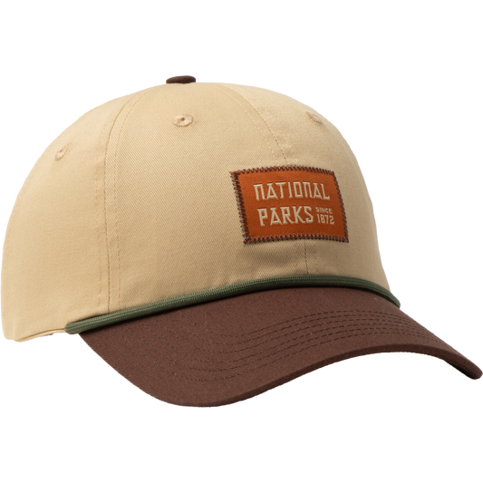 A beige baseball cap with a brown visor, featuring a green piping detail along the bill and a brown leather patch on the front that reads 'NATIONAL PARKS since 1962'.