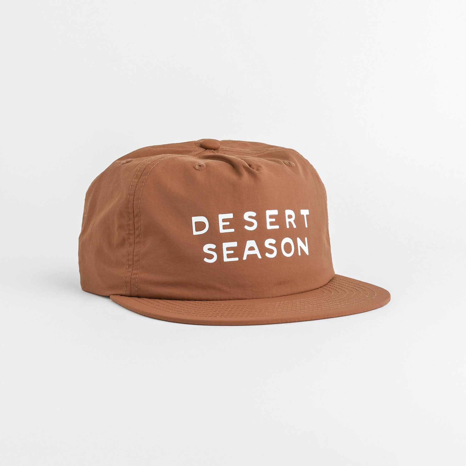 A brown flat-brim cap with 'DESERT SEASON' printed in white text on the front, set against a plain light gray background.