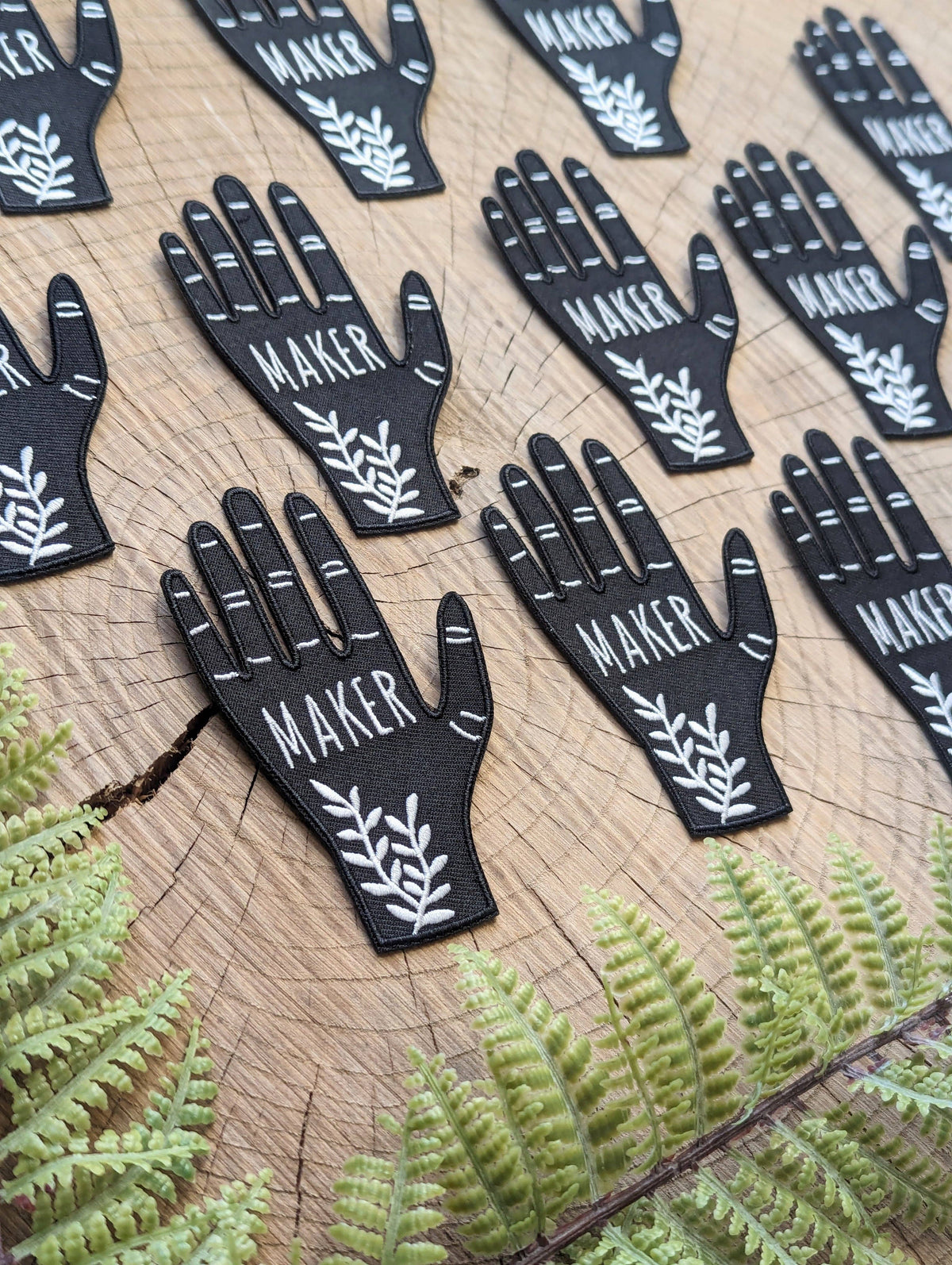 Black hand-shaped patches with 'MAKER' text on a wooden surface with fern leaves.