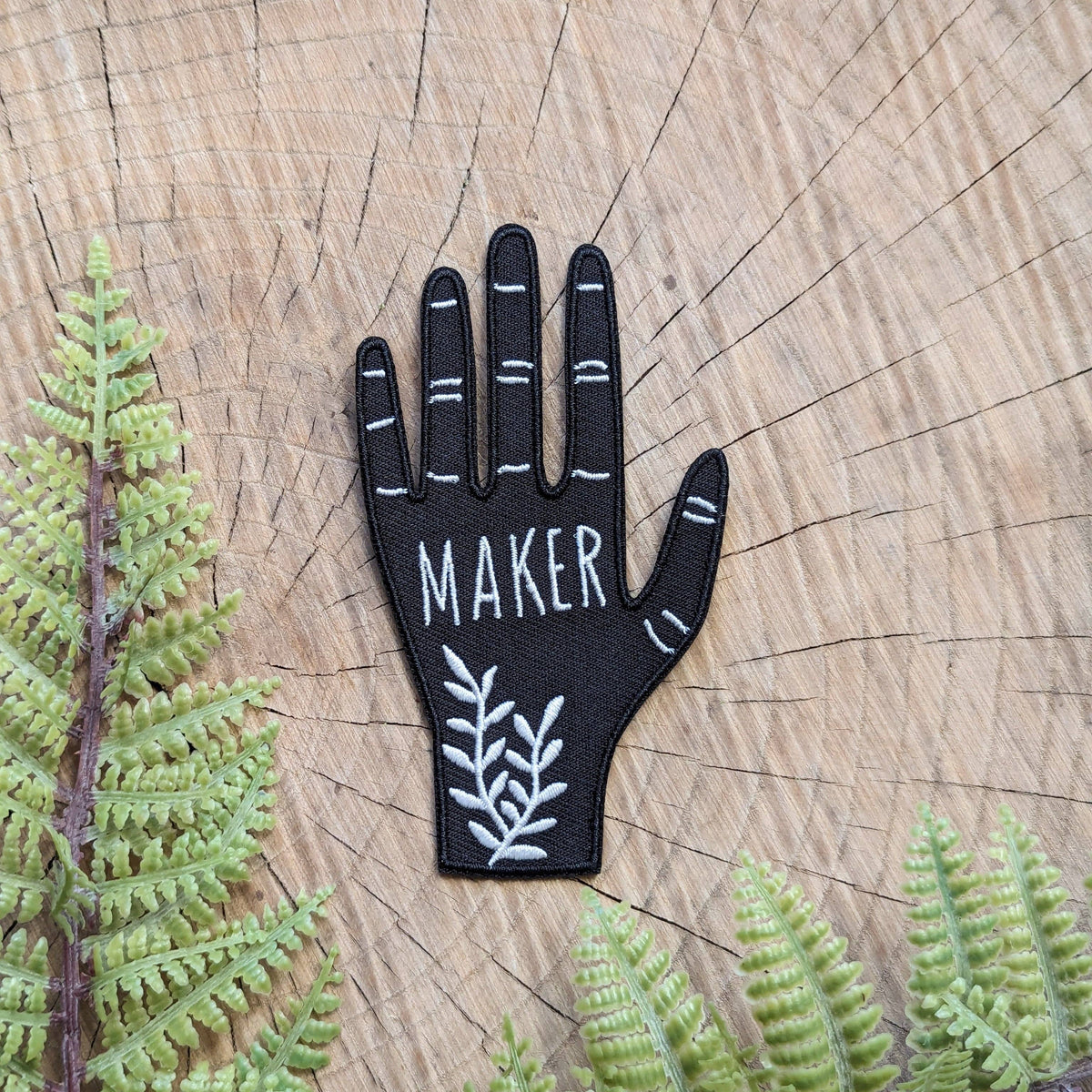 Black hand-shaped patch with 'MAKER' text on a wooden background with ferns.