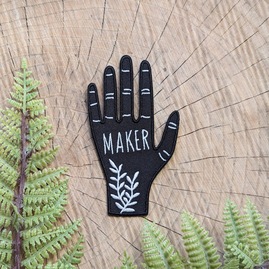 Black hand-shaped patch with 'MAKER' text on a wooden background with ferns.