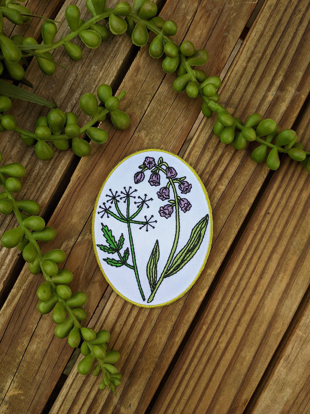 An oval enamel piatch featuring a botanical illustration of purple flowers and green leaves on a white background, bordered in gold. The pin is displayed on a rustic wooden surface decorated with green beaded vines.