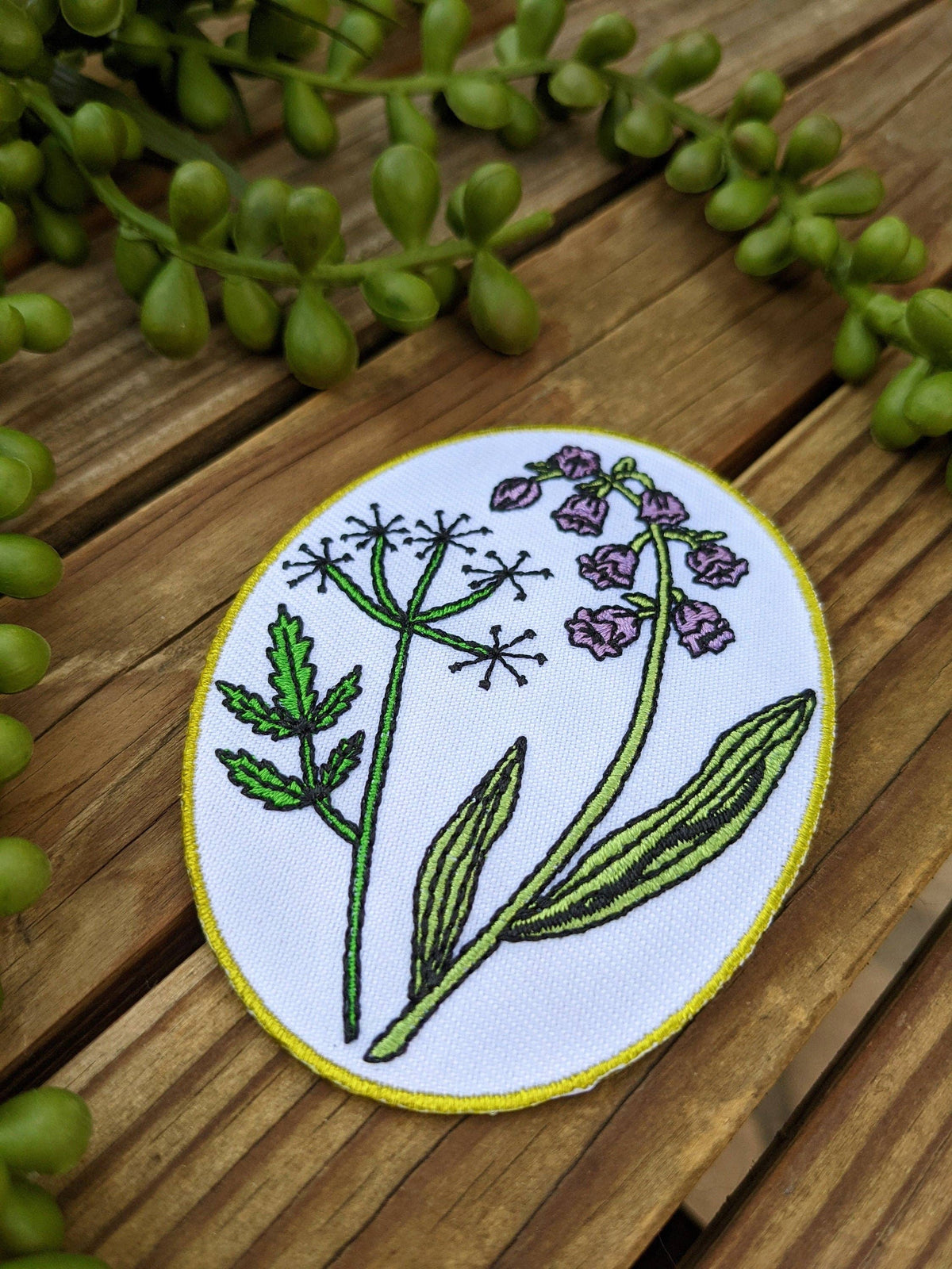 An oval embroidered patch featuring green and purple wildflower designs on a white background with a yellow border, displayed on a wooden surface with green trailing leaves.