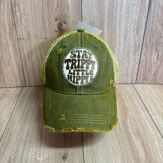 An olive green distressed trucker cap with a circular embroidered patch reading 'STAY TRIPPY LITTLE HIPPIE' on the front, set against a light wood plank background.