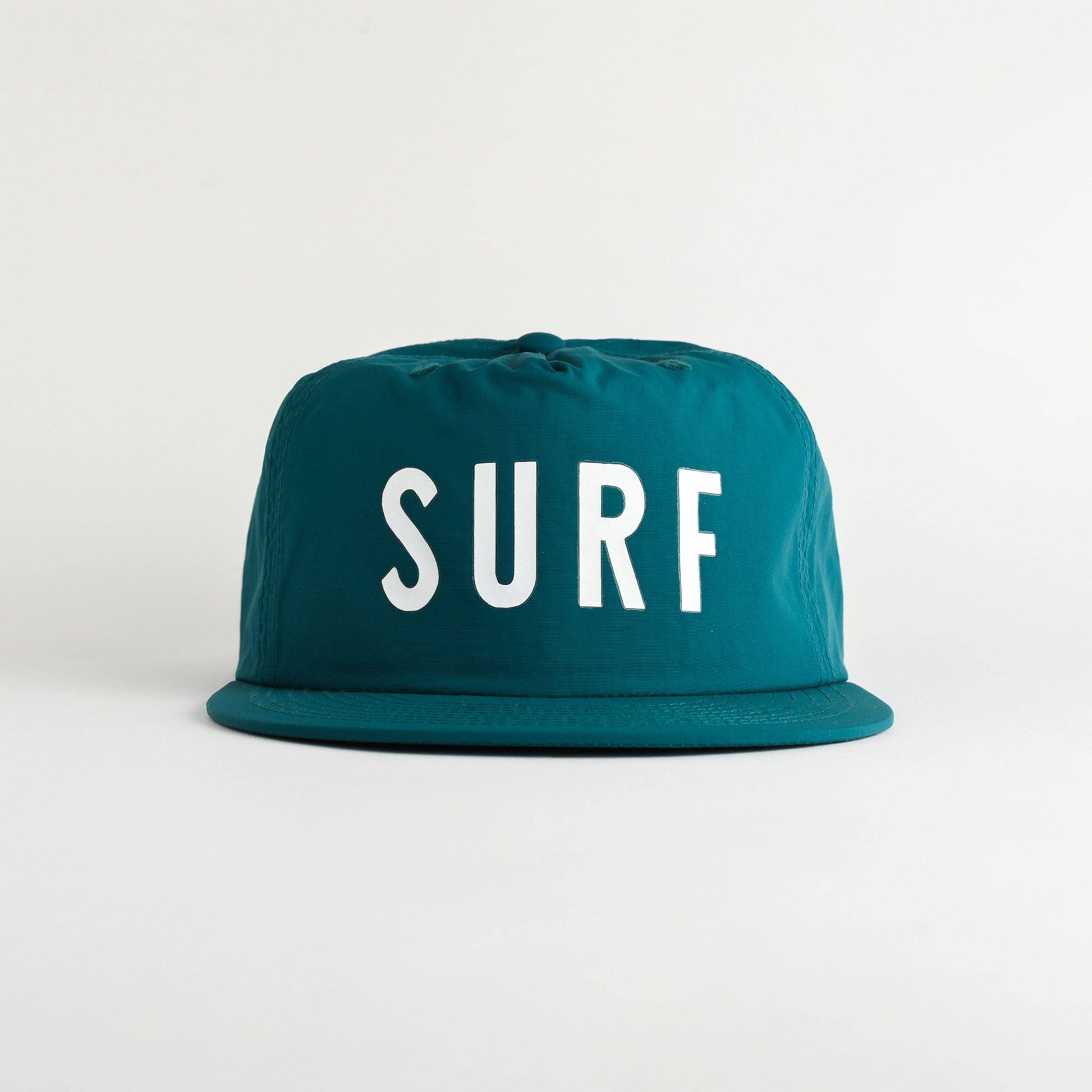 A teal blue flat-brim cap with the word 'SURF' printed in bold white capital letters on the front, set against a plain light gray background.