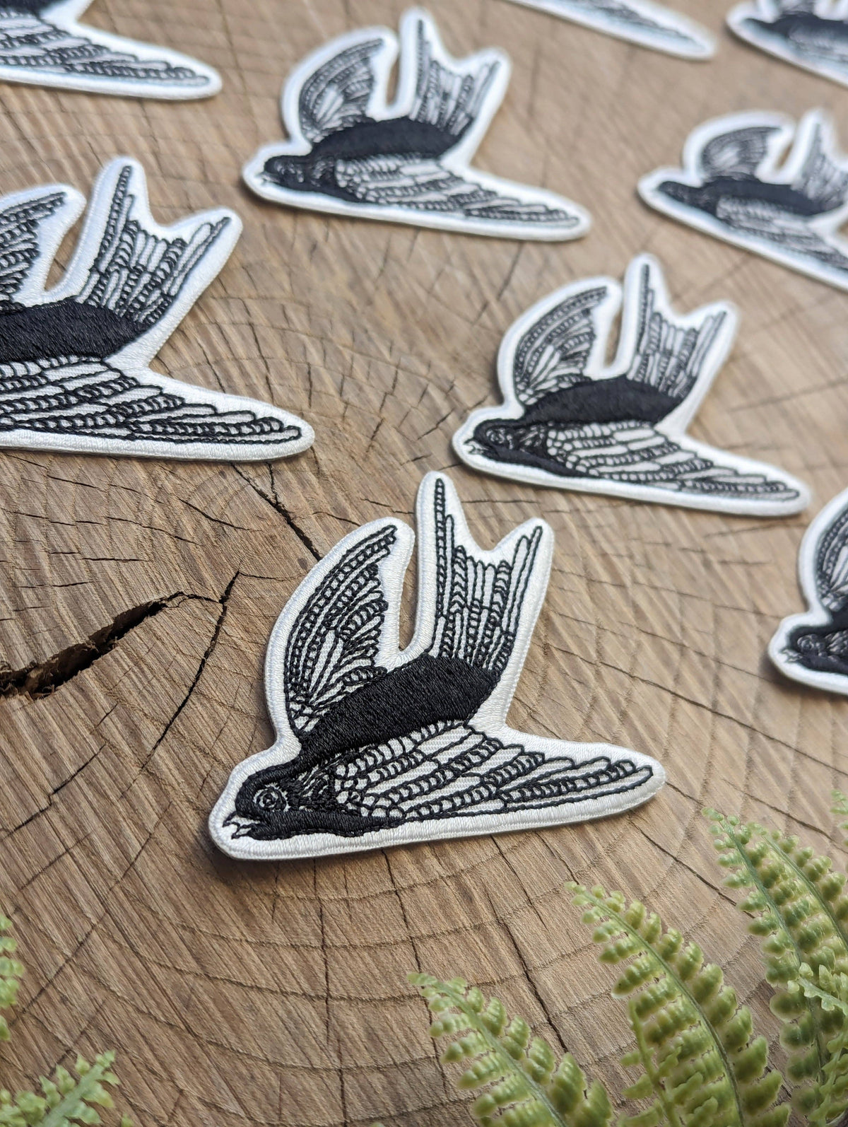 Black and white embroidered swallow patch with a white border, displayed on a rustic wood slice background with green fern leaves in the foreground.